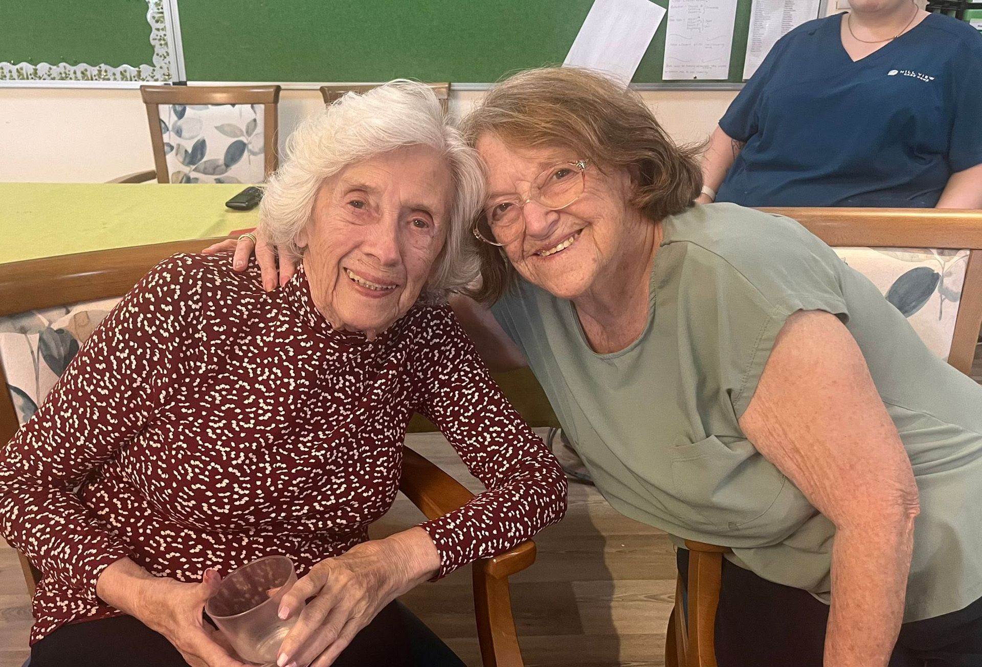 Two smiling women lean together at a table in a bright room, with a person in blue seated behind them.