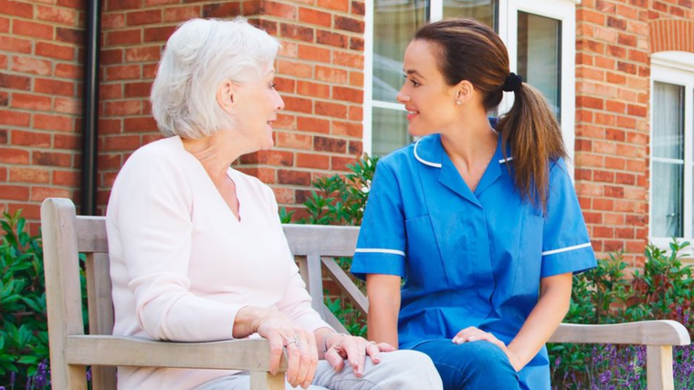 Two women talking on a bench outside a brick building, one in white and one in blue uniforms.