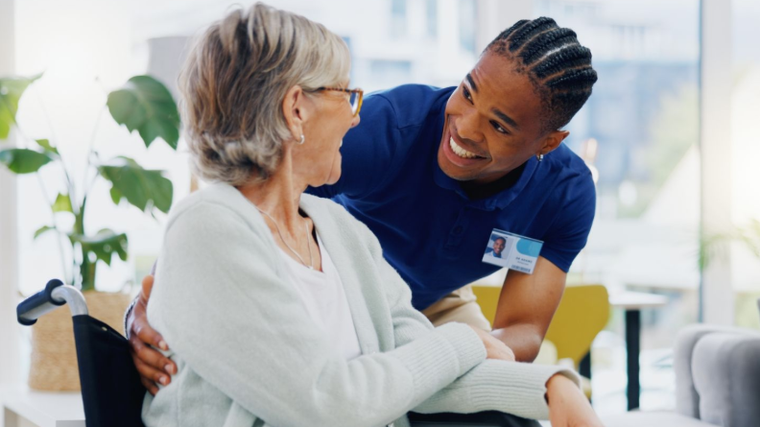 Nurse in blue smiling and speaking with an older patient in a bright clinic waiting area