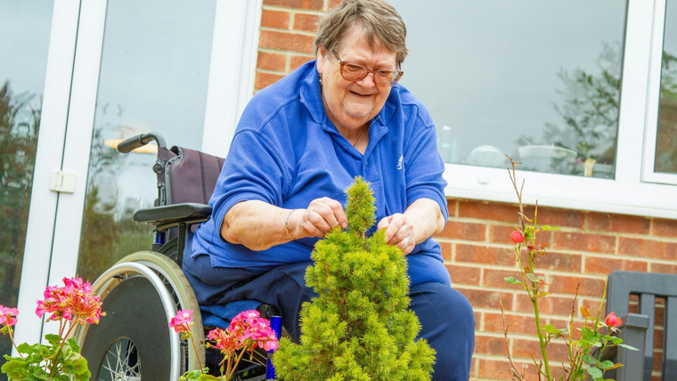 Person in a blue shirt tending a potted plant beside a wheelchair outside a brick house