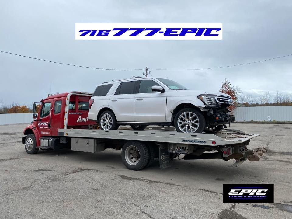 Red tow truck carrying a wrecked white SUV, with TIB EPIC and EPIC logos displayed above and below.
