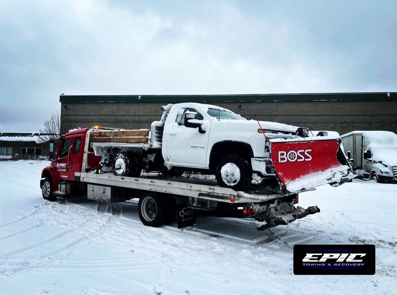 Tow truck carrying a white pickup and red sled in a snowy lot with a building behind it