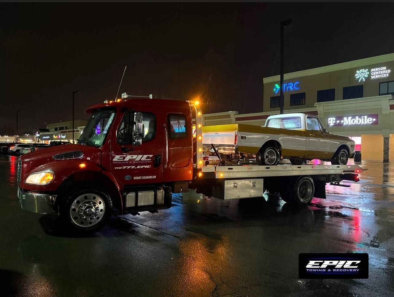 Red tow truck hauling a beige pickup on a wet street at night in front of lit storefronts.