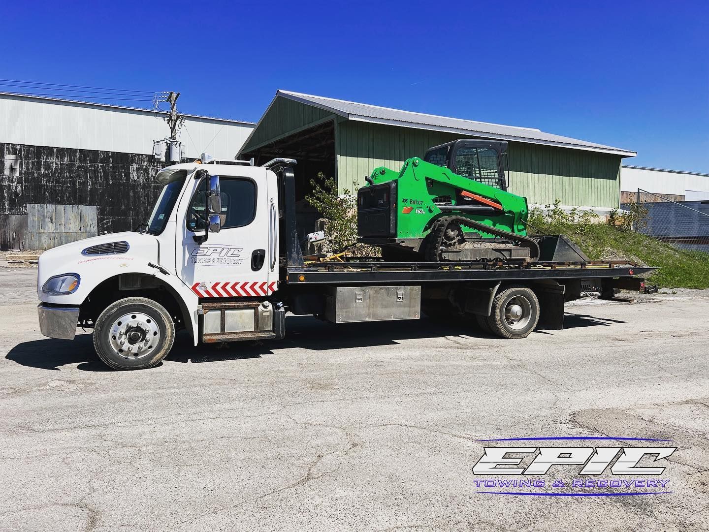 White flatbed truck carrying a green machine parked in a gravel lot by a metal building