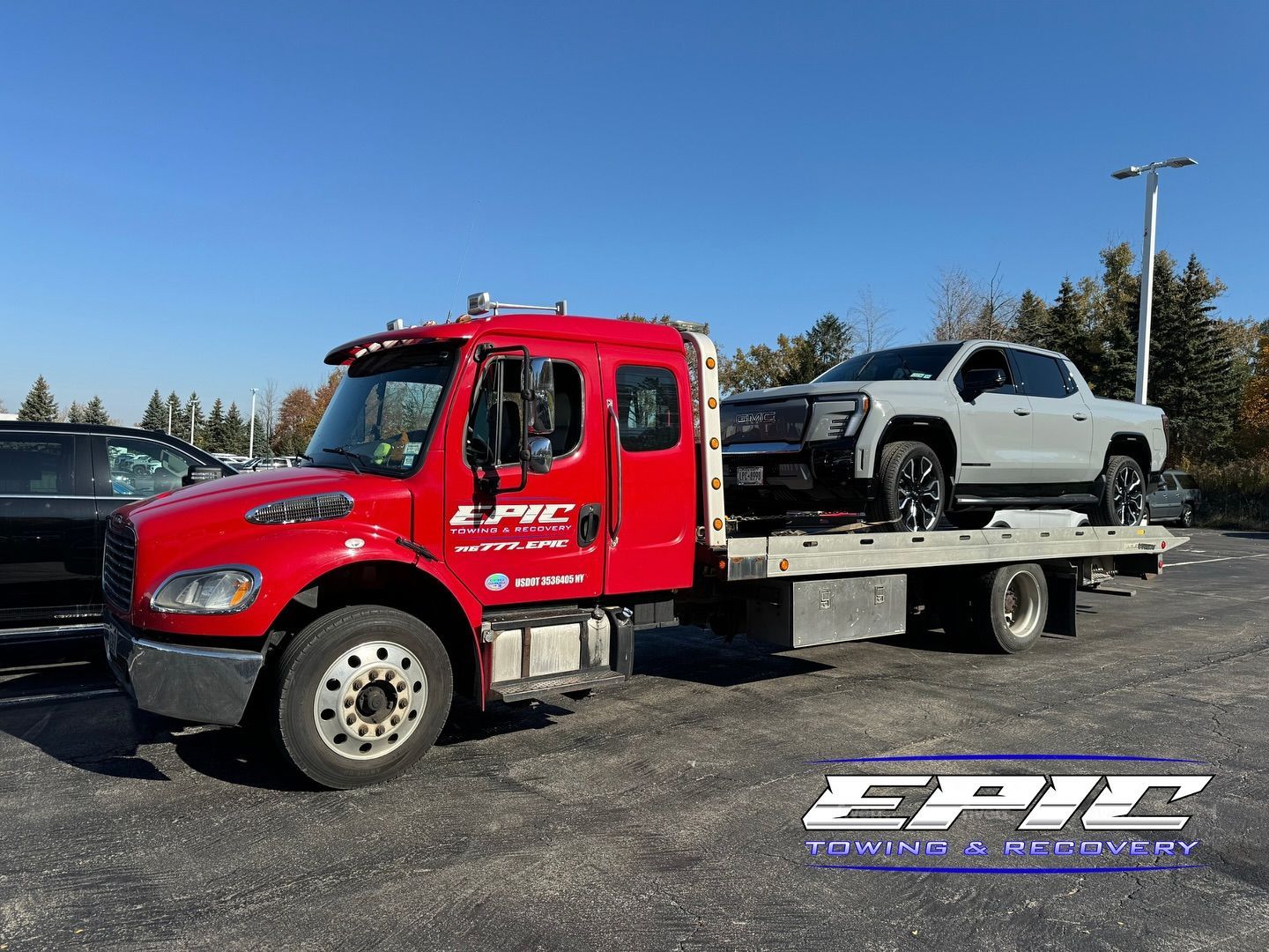 Red tow truck carrying a silver pickup on a flatbed in a parking lot under a clear blue sky