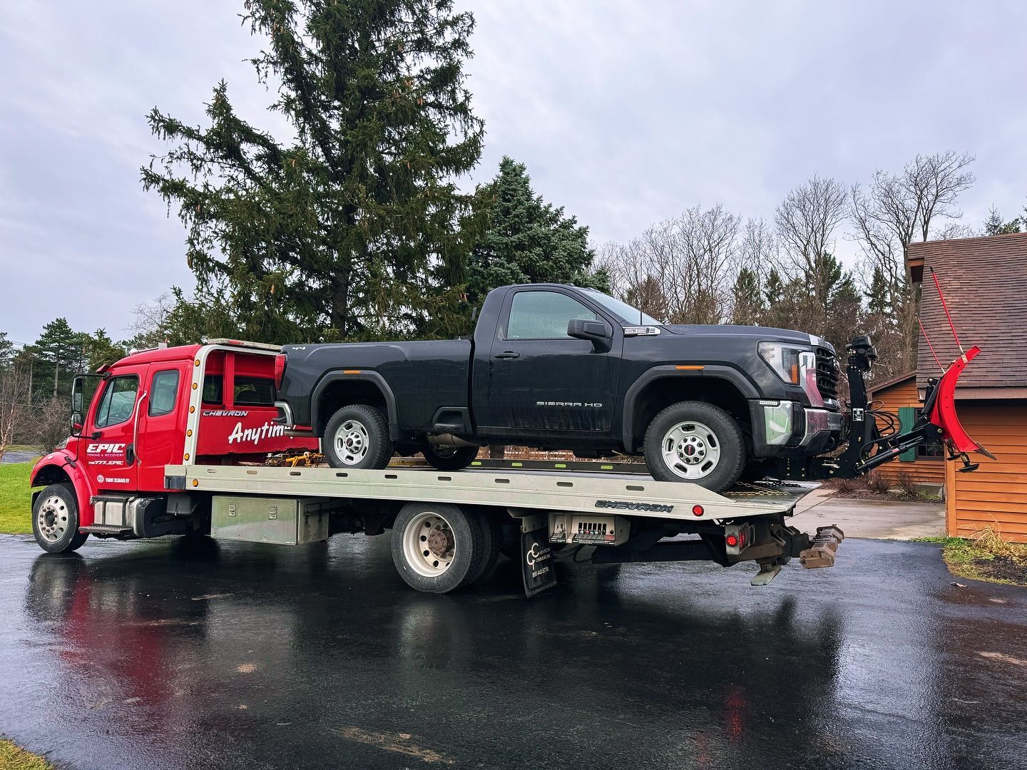 Red tow truck hauling a black pickup on a flatbed in a wet parking lot.