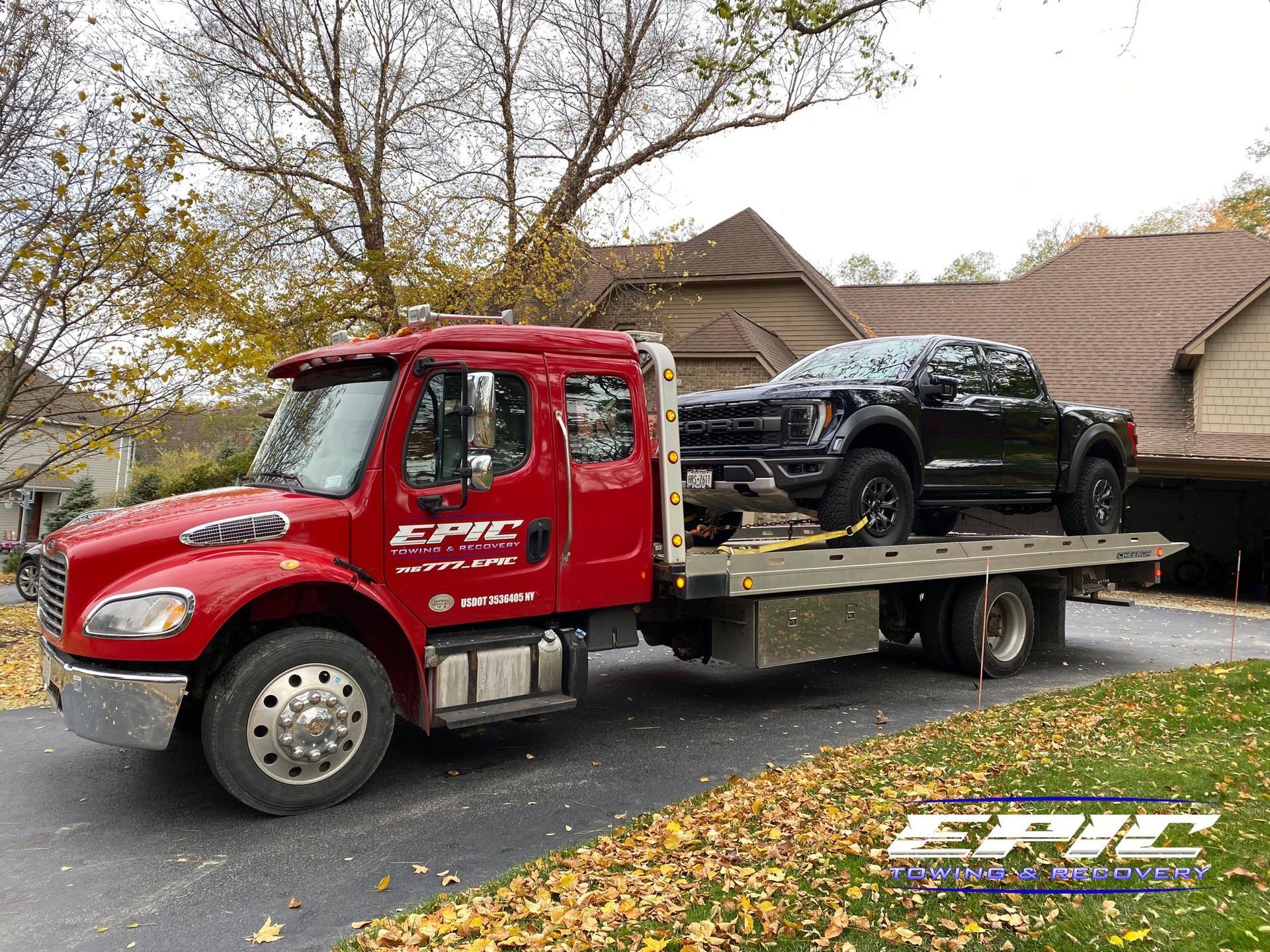 Red EPIC tow truck hauling a black car on a flatbed in a suburban driveway with autumn leaves.