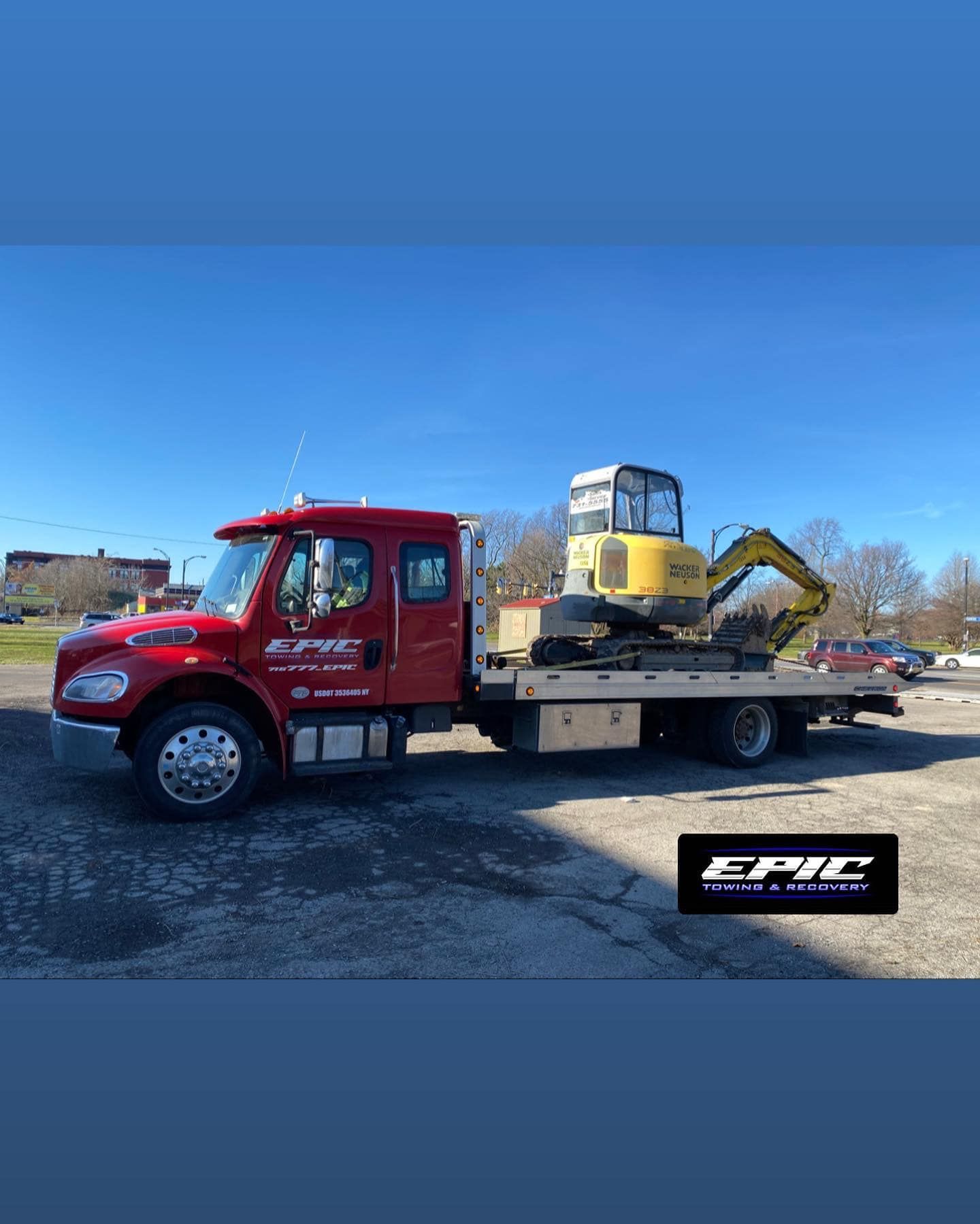 Red tow truck hauling a yellow excavator on a flatbed in a parking lot under a blue sky