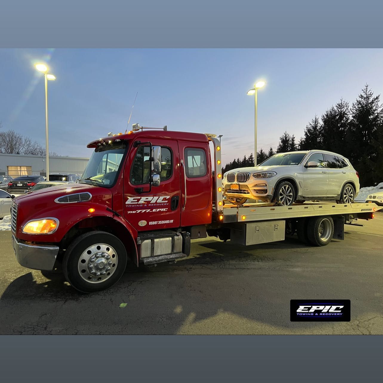 Red flatbed tow truck carrying a silver SUV in a parking lot at dusk