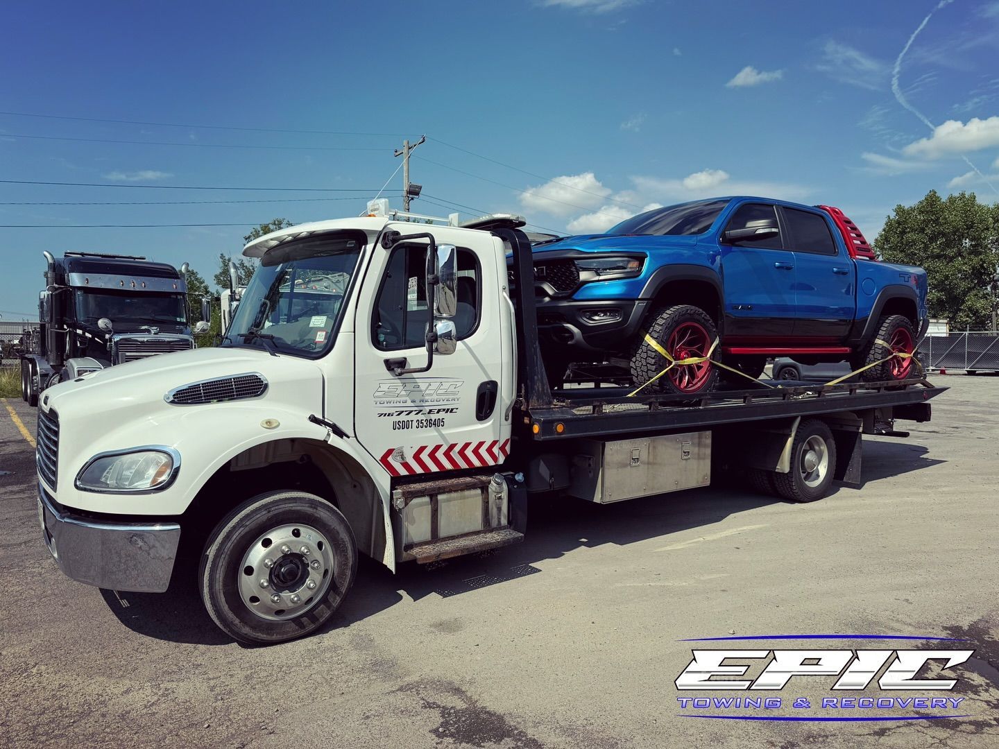 Tow truck carrying a blue pickup on a flatbed under a sunny sky.