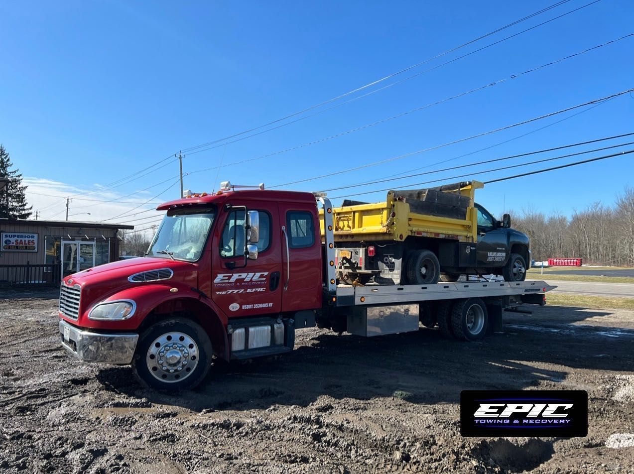 Red tow truck hauling yellow equipment on a muddy lot under a clear blue sky
