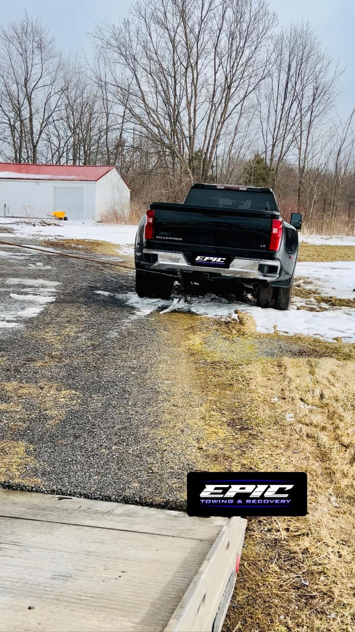 Black pickup truck on a gravel driveway beside snow patches, with trees and a small building in the background.