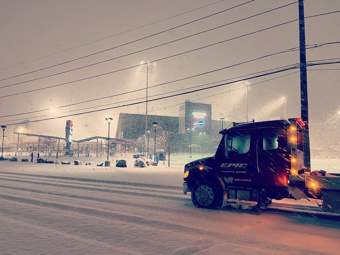 Snowplow clearing a snowy road at night near an illuminated stadium or arena