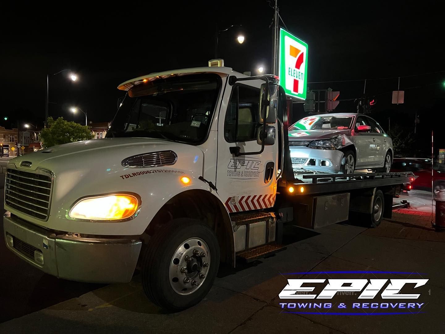 Tow truck towing a car at night outside a 7-Eleven parking lot