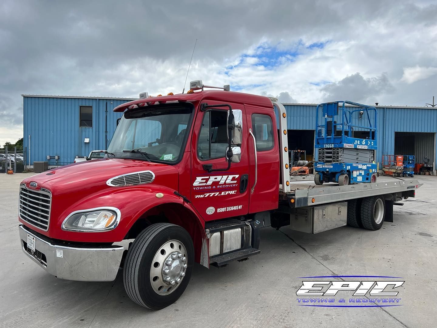 Red flatbed tow truck parked in an industrial yard with blue buildings and cloudy sky.