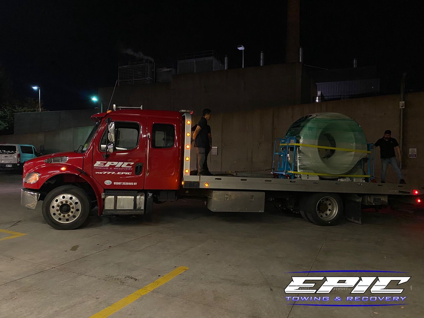 Red tow truck hauling a large cable reel on a flatbed in a dimly lit parking lot at night