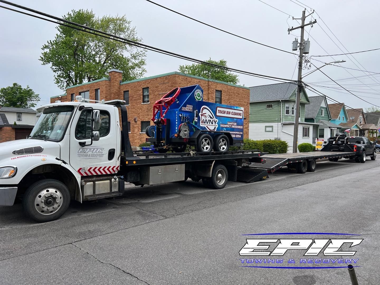 Tow truck hauling a blue race car trailer down a residential street, with parked cars and houses nearby.