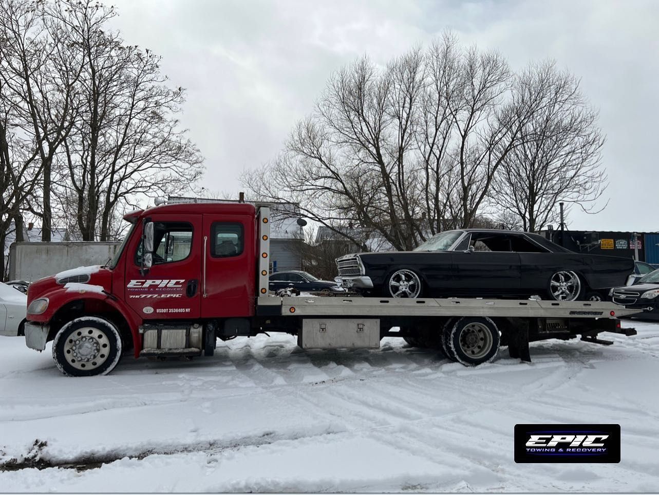 Red tow truck carrying a black car on a snowy road with trees in the background