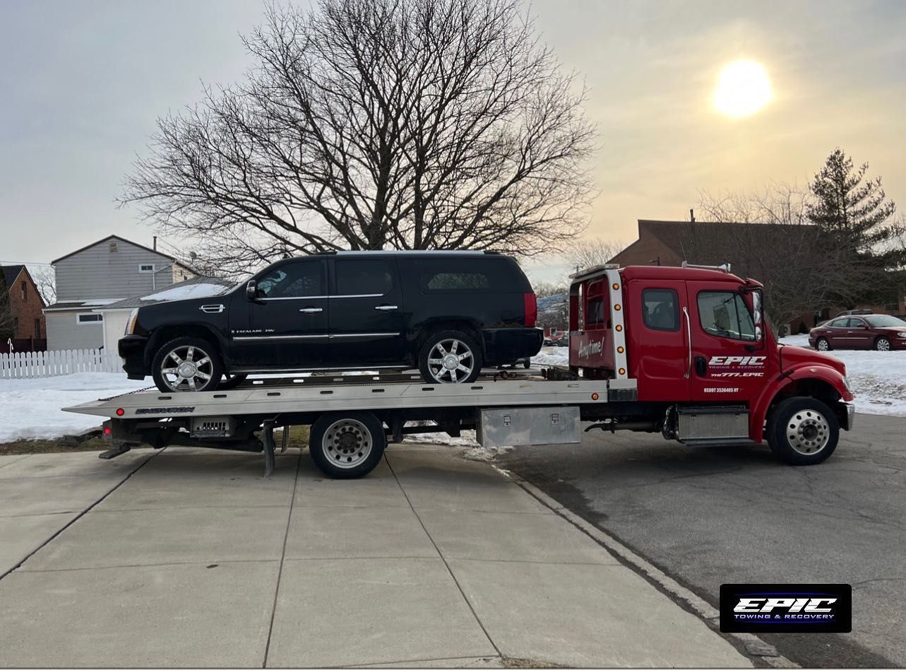 Red tow truck carrying a black SUV on a flatbed in a snowy driveway at sunset
