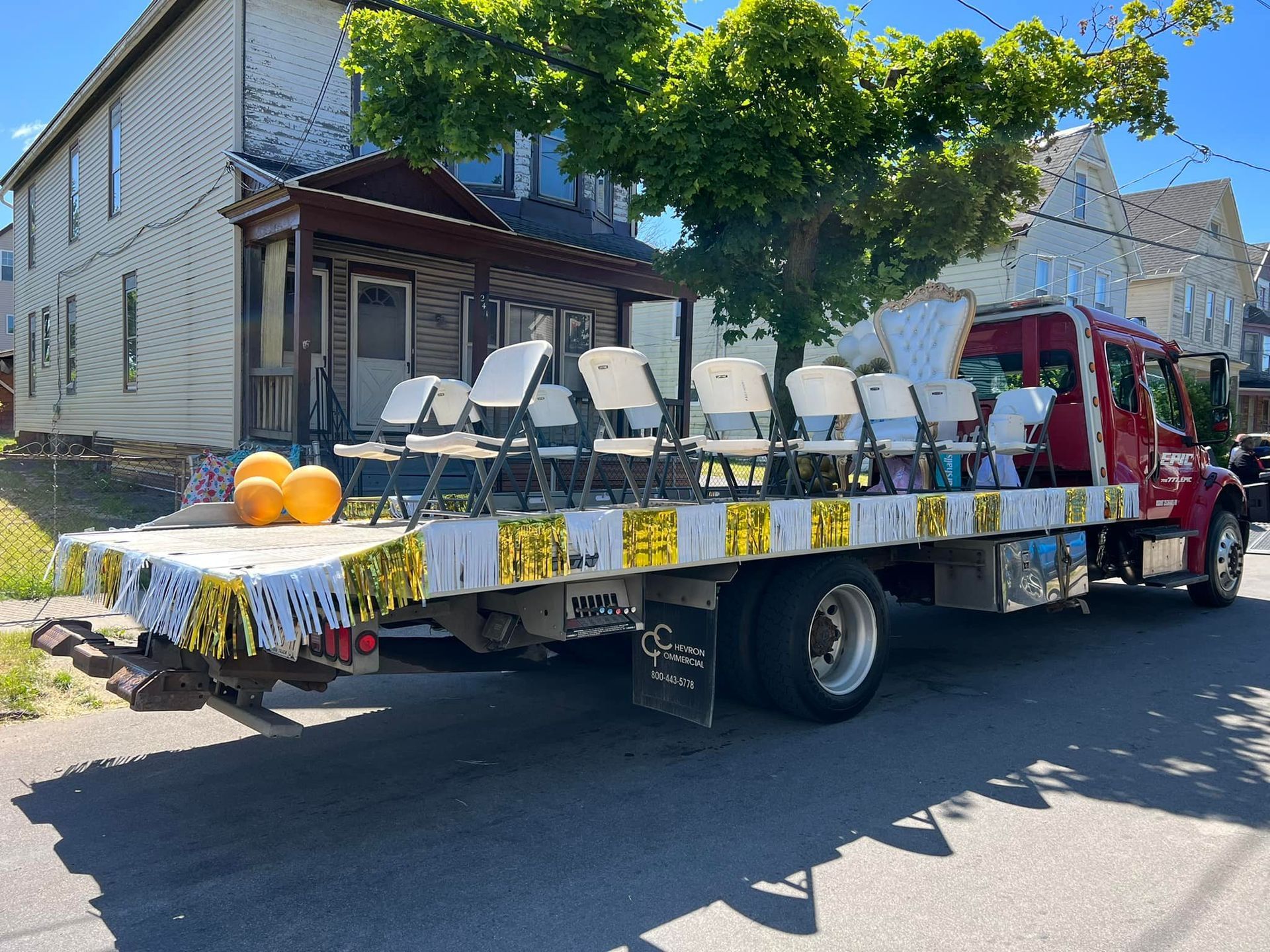 Float truck with white chairs and table decorations, parked on a sunny street during a parade.