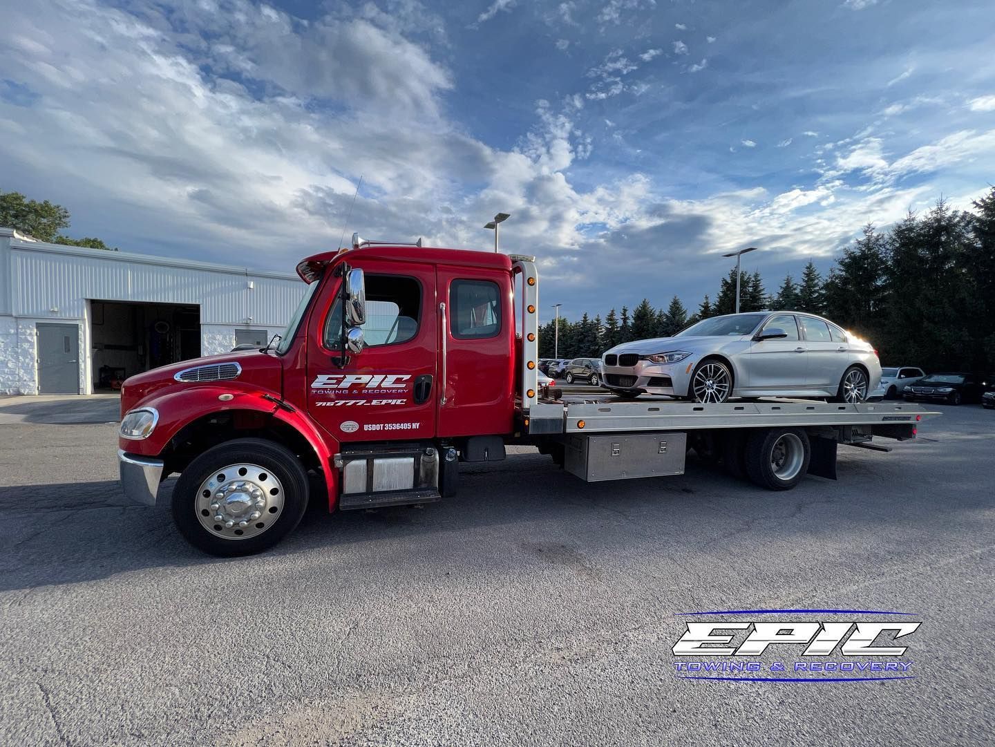 Red flatbed tow truck carrying a silver sedan in a gravel lot under a cloudy sky