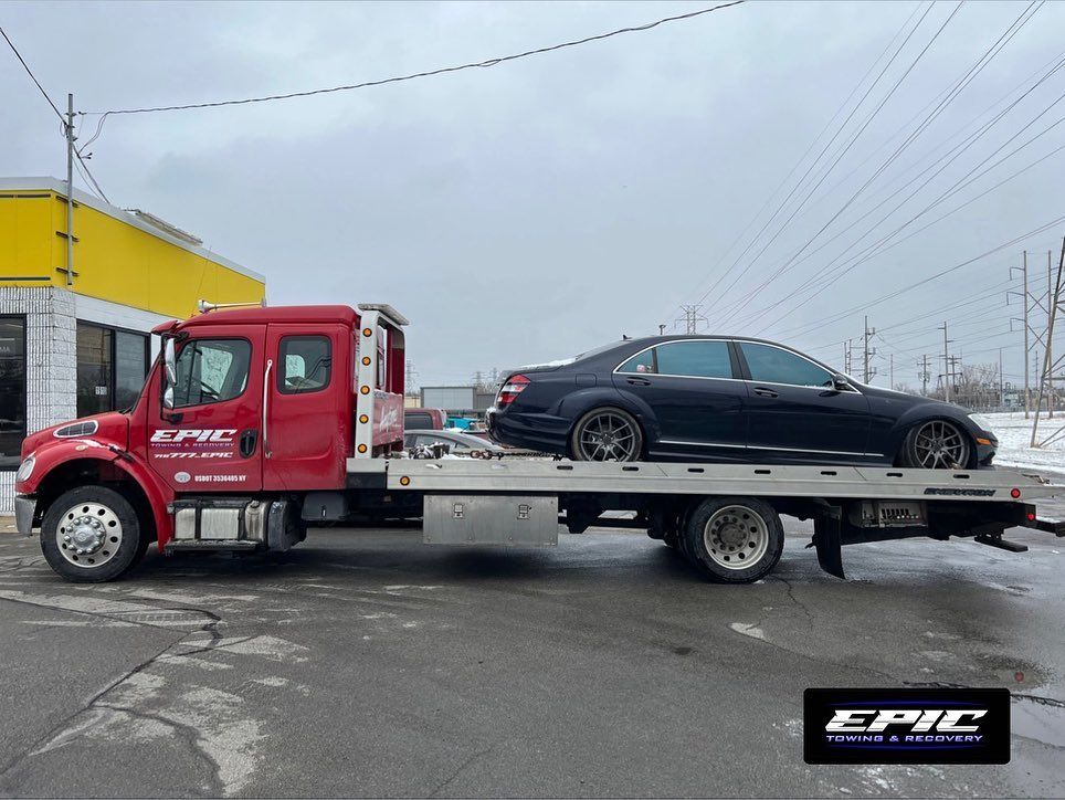 Red flatbed tow truck carrying a black sedan on a roadside under cloudy skies