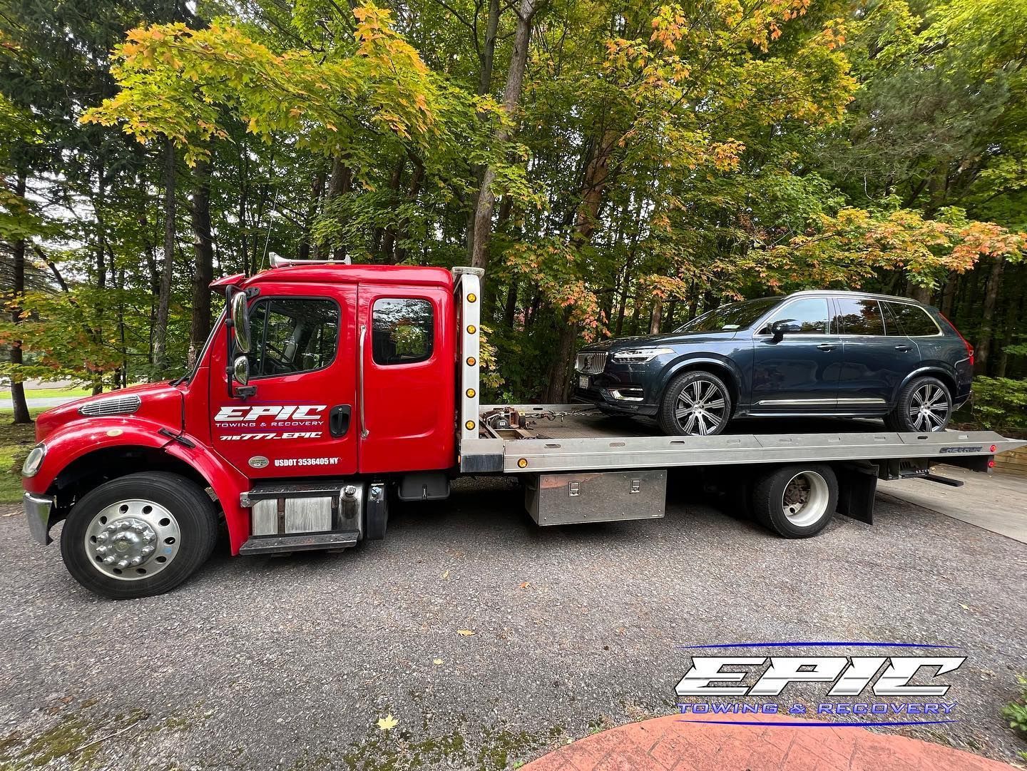 Red flatbed tow truck carrying a black SUV on a gravel lot with trees in the background