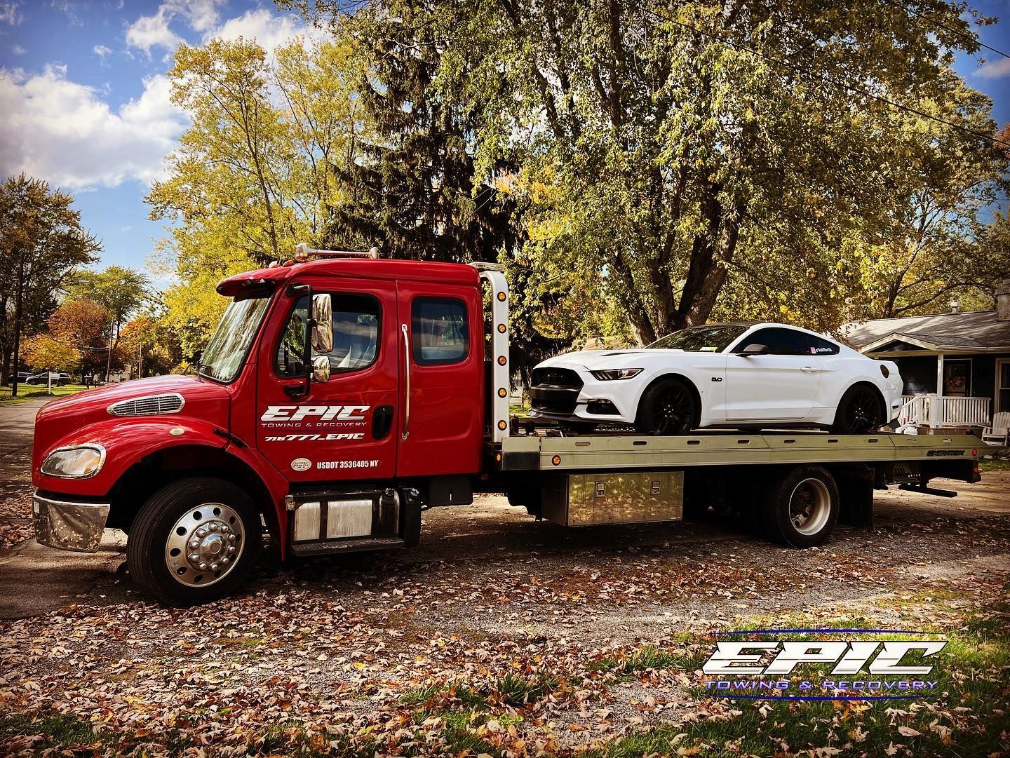 Red tow truck carrying a white sports car on a flatbed in an autumn park.