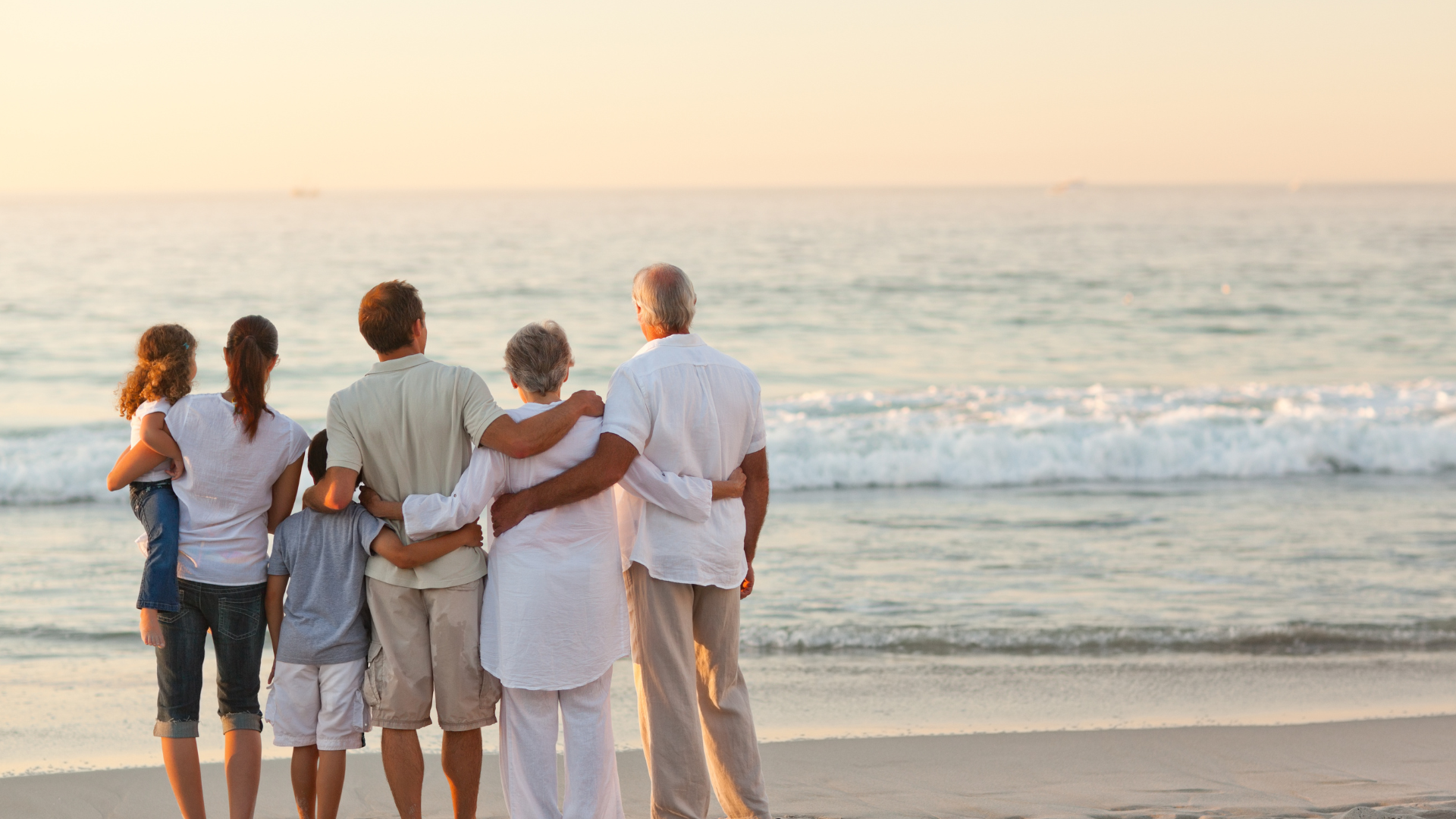 Une famille de sept personnes debout sur une plage, les bras les uns autour des autres, regardant l'océan pendant un coucher de soleil.
