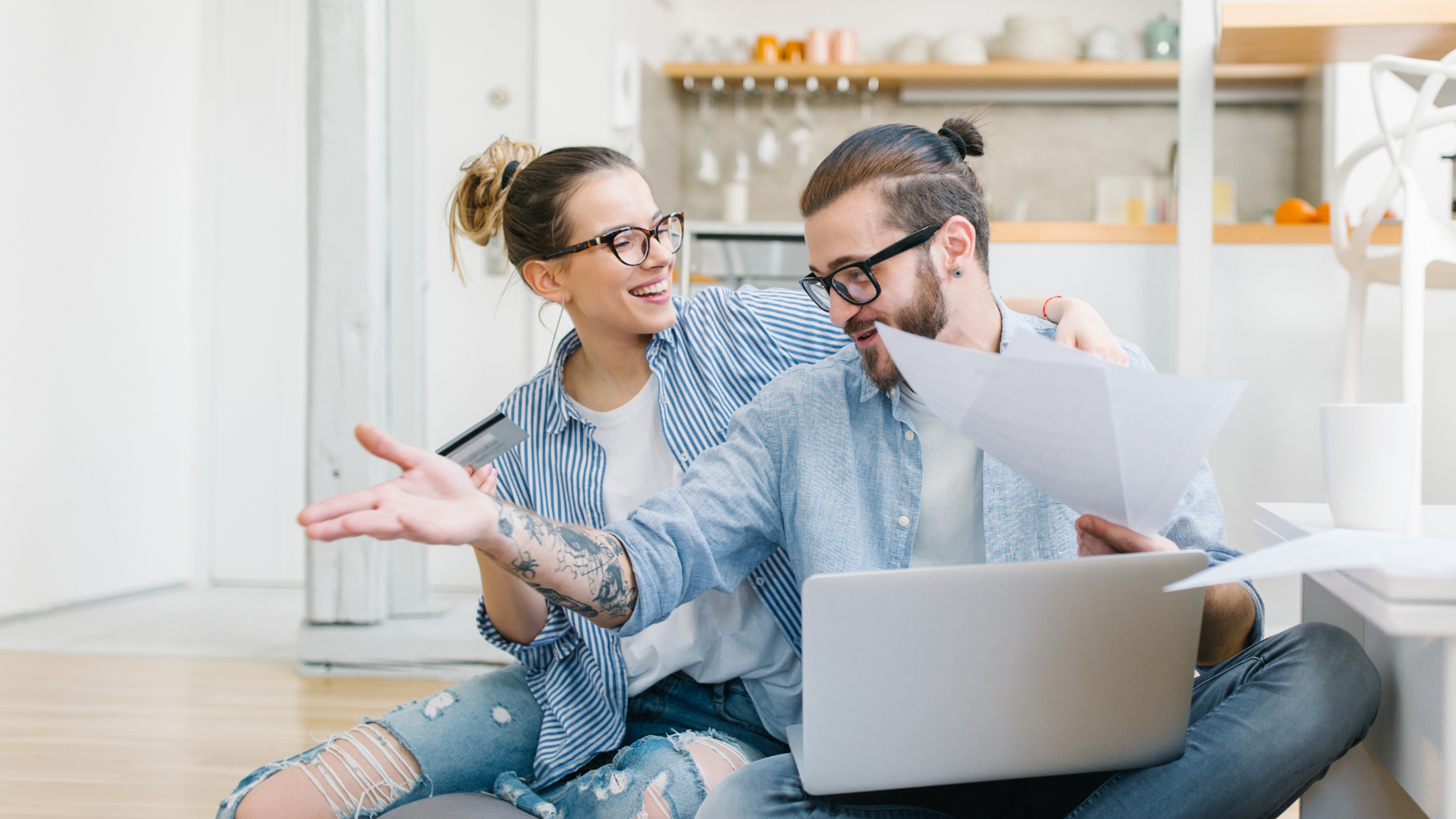 Un couple est assis par terre avec un ordinateur portable, des papiers et une carte de crédit. Ils consultent leurs papiers avec un air heureux, dans un intérieur moderne.