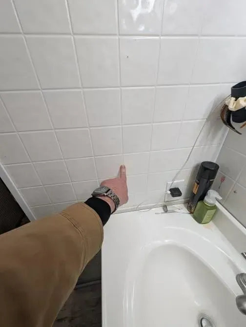 A person is pointing at a tiled wall in a bathroom next to a sink.