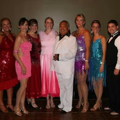 Group of people in colorful formal outfits posing onstage, with one person in a white suit at center