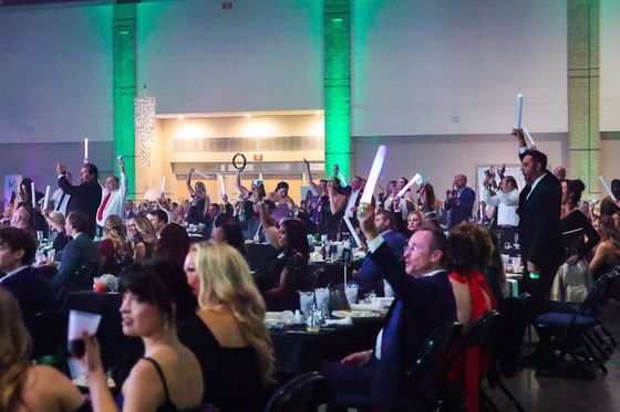 Crowd at a banquet applauding in a large hall with green-lit columns and tables set with drinks