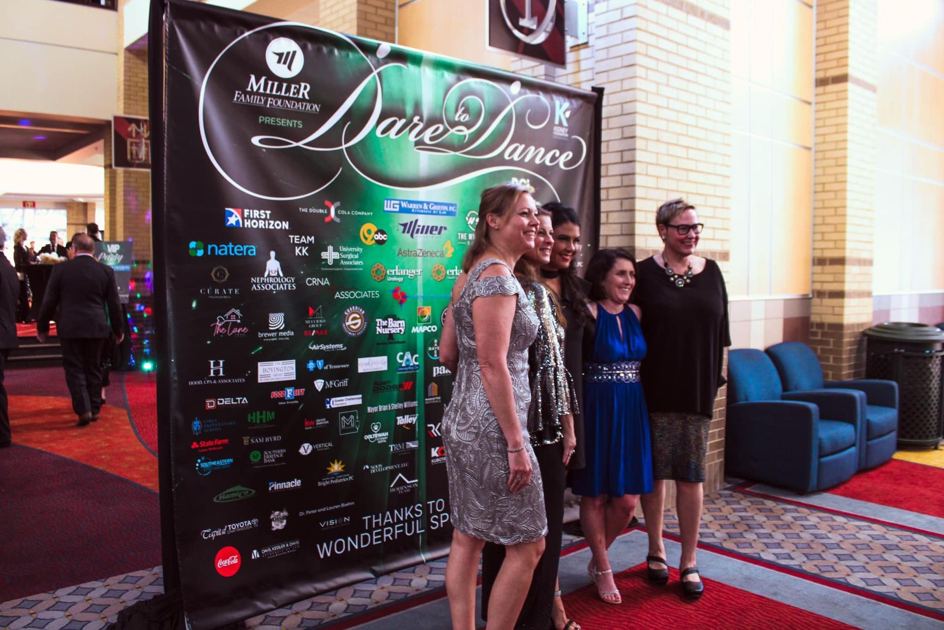 Three people posing on a red carpet beside a sponsor backdrop at a dance event.