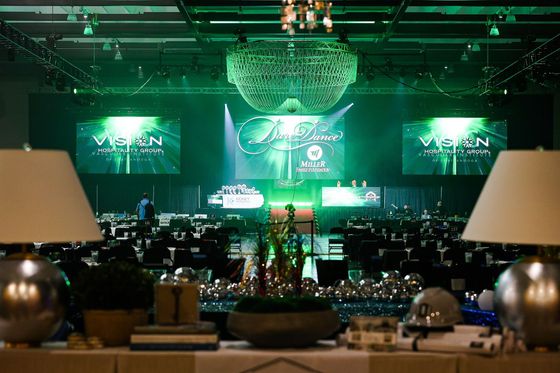 Green-lit conference hall with stage screens and chandeliers, viewed from a dining table.