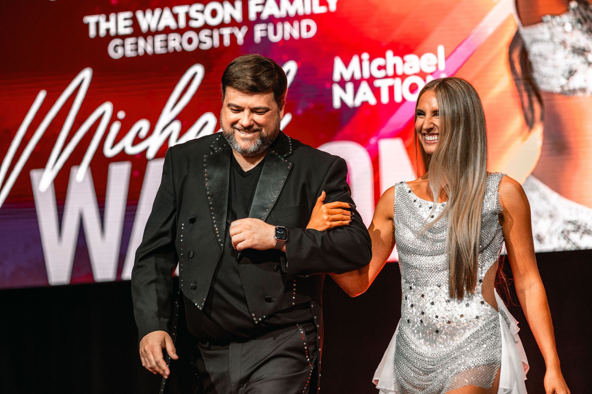 Two people holding hands on a red carpet in front of a Michael event backdrop