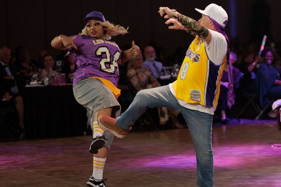 Two dancers in purple and yellow jerseys kick in unison on a lit stage