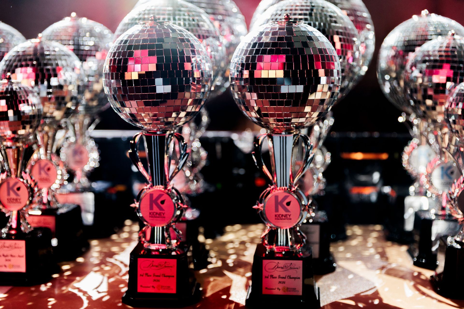 Rows of shiny silver trophies with pink bases and plaques on a stage under confetti lights