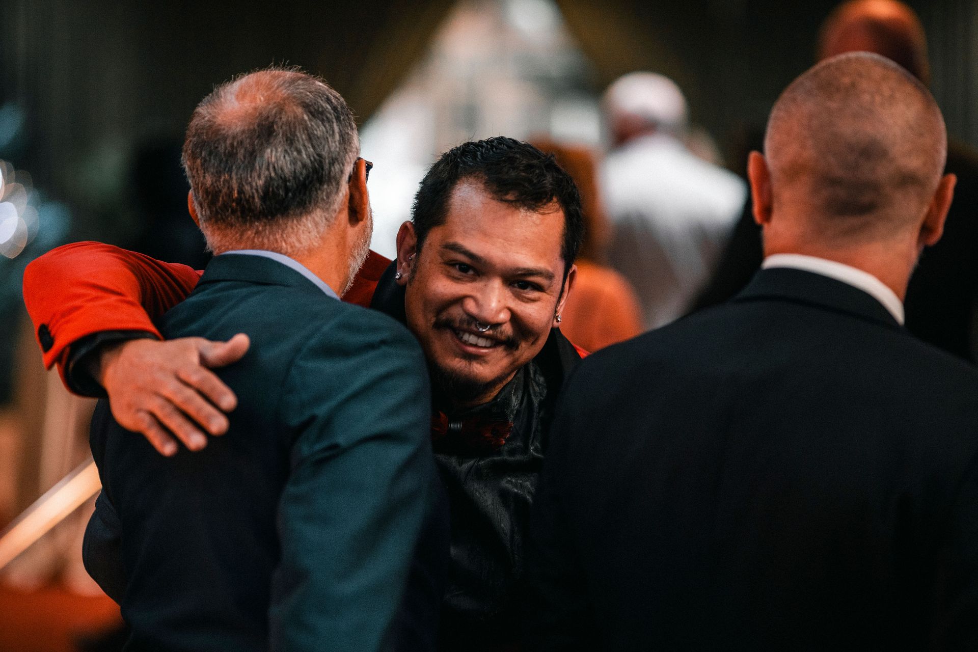 Two men embracing warmly in a crowded indoor event setting, with another man in a dark suit in the foreground