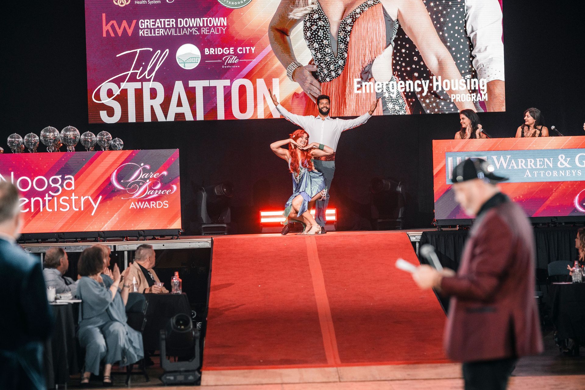 Dancer on red-carpeted stage at pageant event, with sponsor banners and audience in front