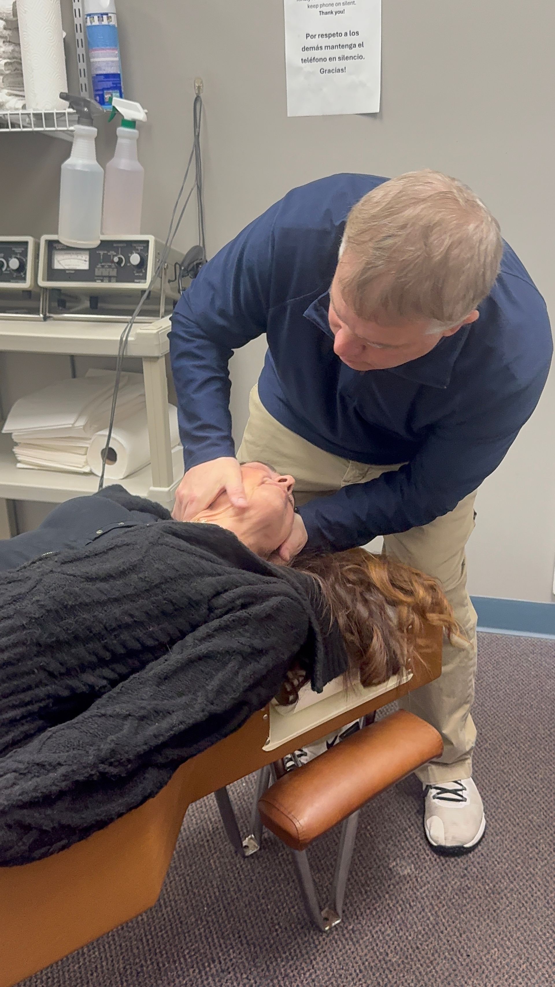 A person receiving a neck adjustment from a chiropractor. The person lies on a padded table. The chiropractor is in a blue shirt.