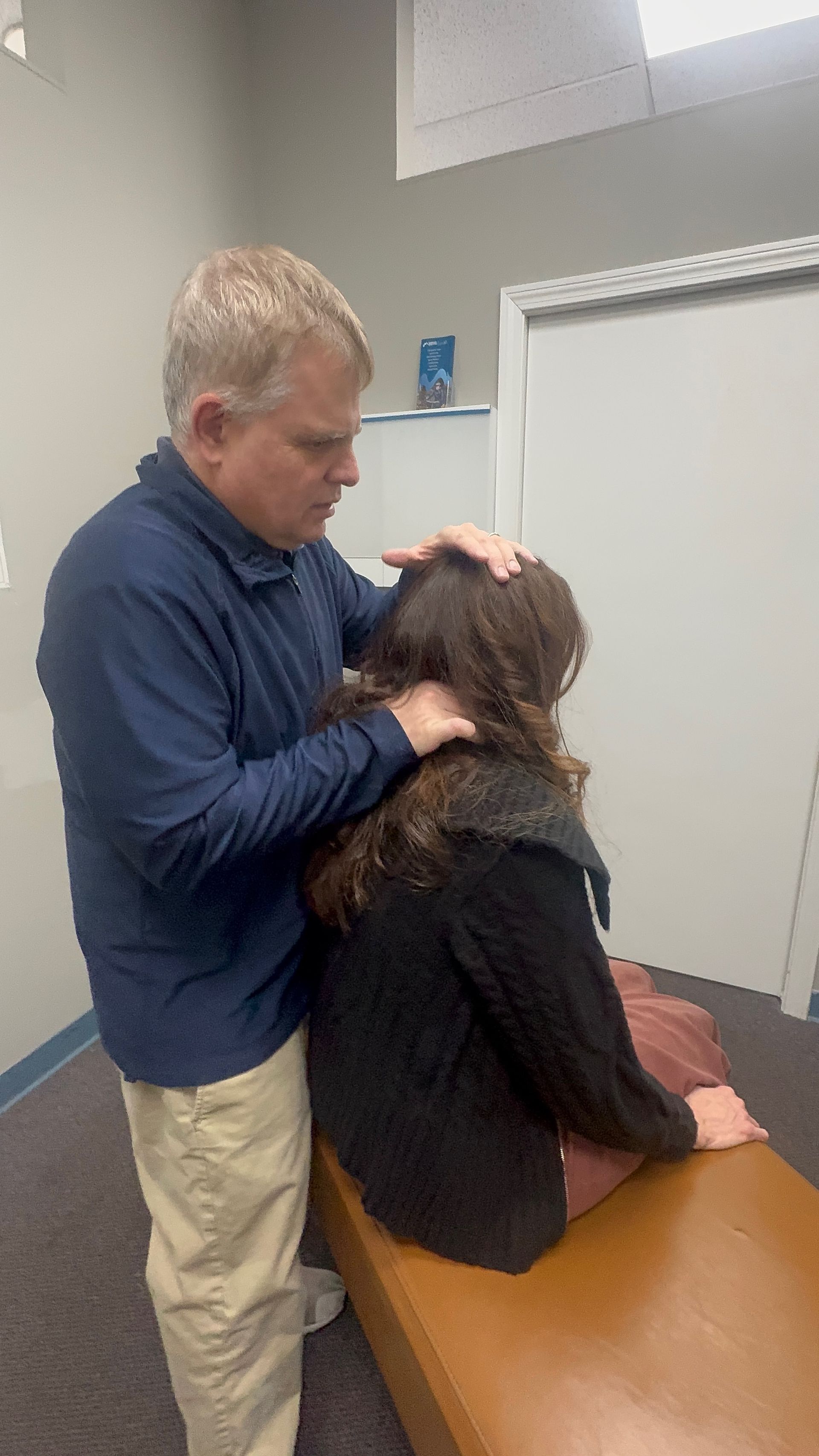 A person in a blue shirt adjusts another person's neck. They're in an office setting.