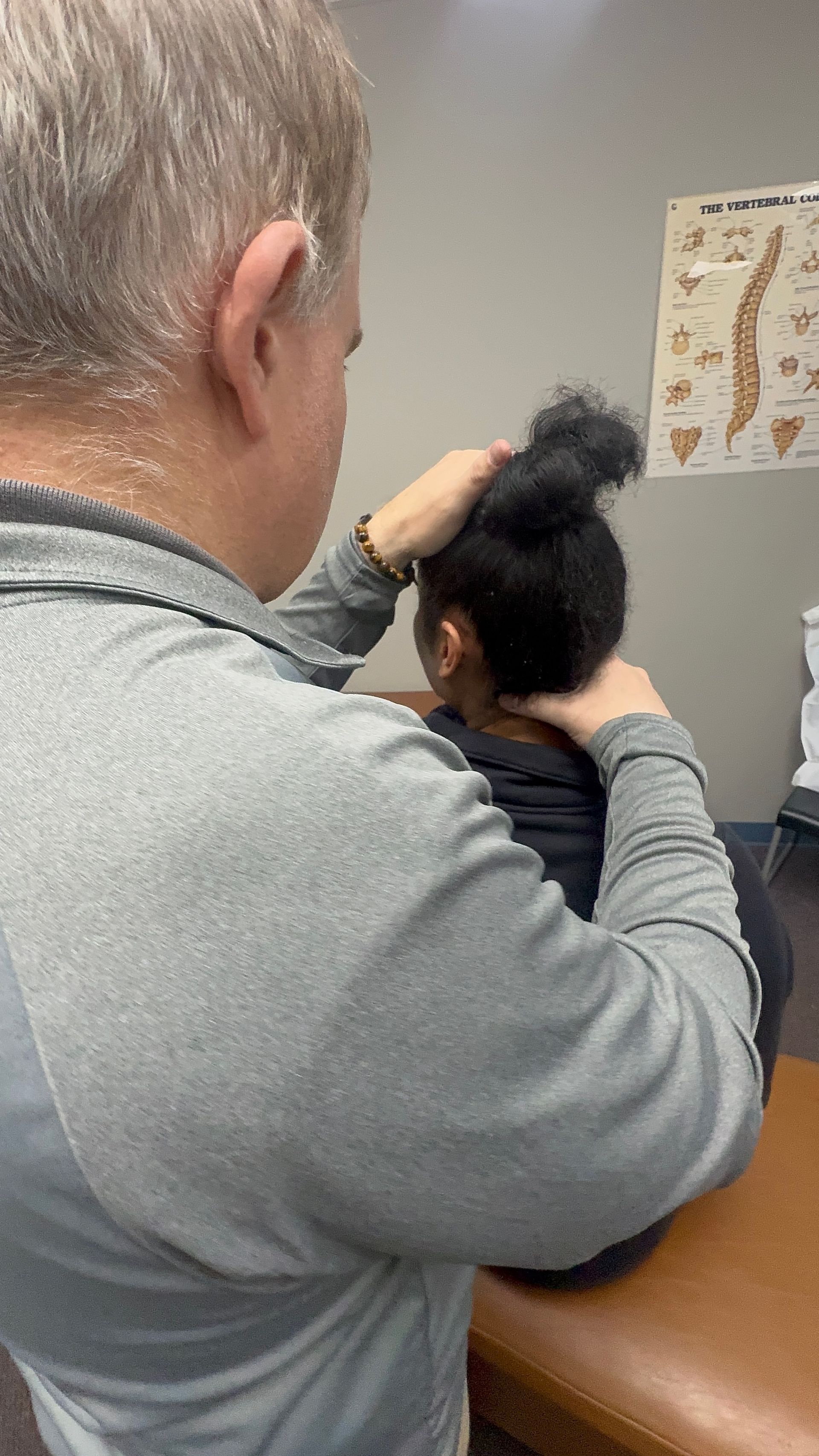 A person examines another person's neck. They sit on a brown bench indoors near a poster.
