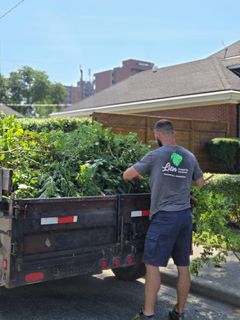 Man spraying plants with hose in backyard next to brick building and covered shrub.