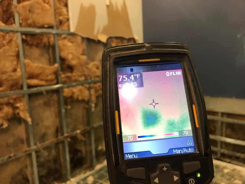 Thermal camera displaying temperature readings; wall with insulation visible behind a wire mesh.