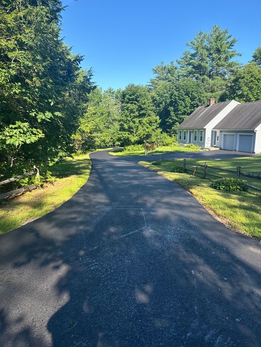 Asphalt driveway curves toward a house with a gray roof, surrounded by green trees and grass, on a sunny day.