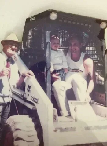Man, child, and woman in a Bobcat. Man wears hat and holds item. Child is in the middle. Woman smiles and holds a bottle.