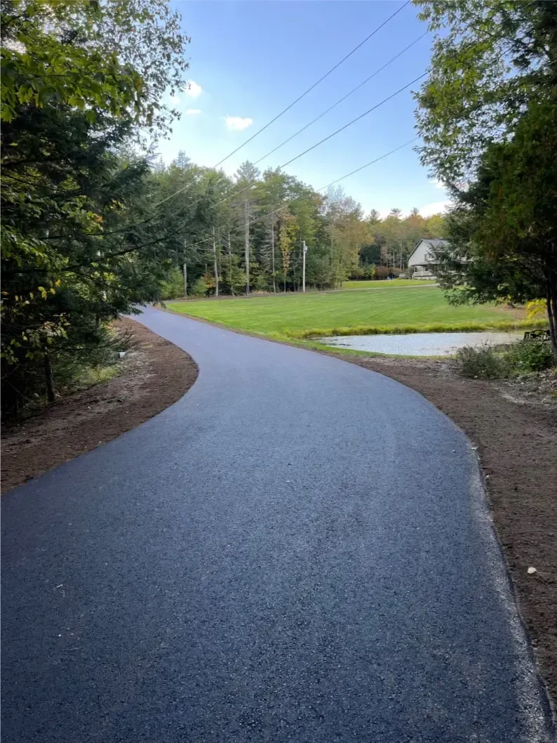 Paved road curves through a green, wooded area. A grassy lawn with a pond and house are in the distance.