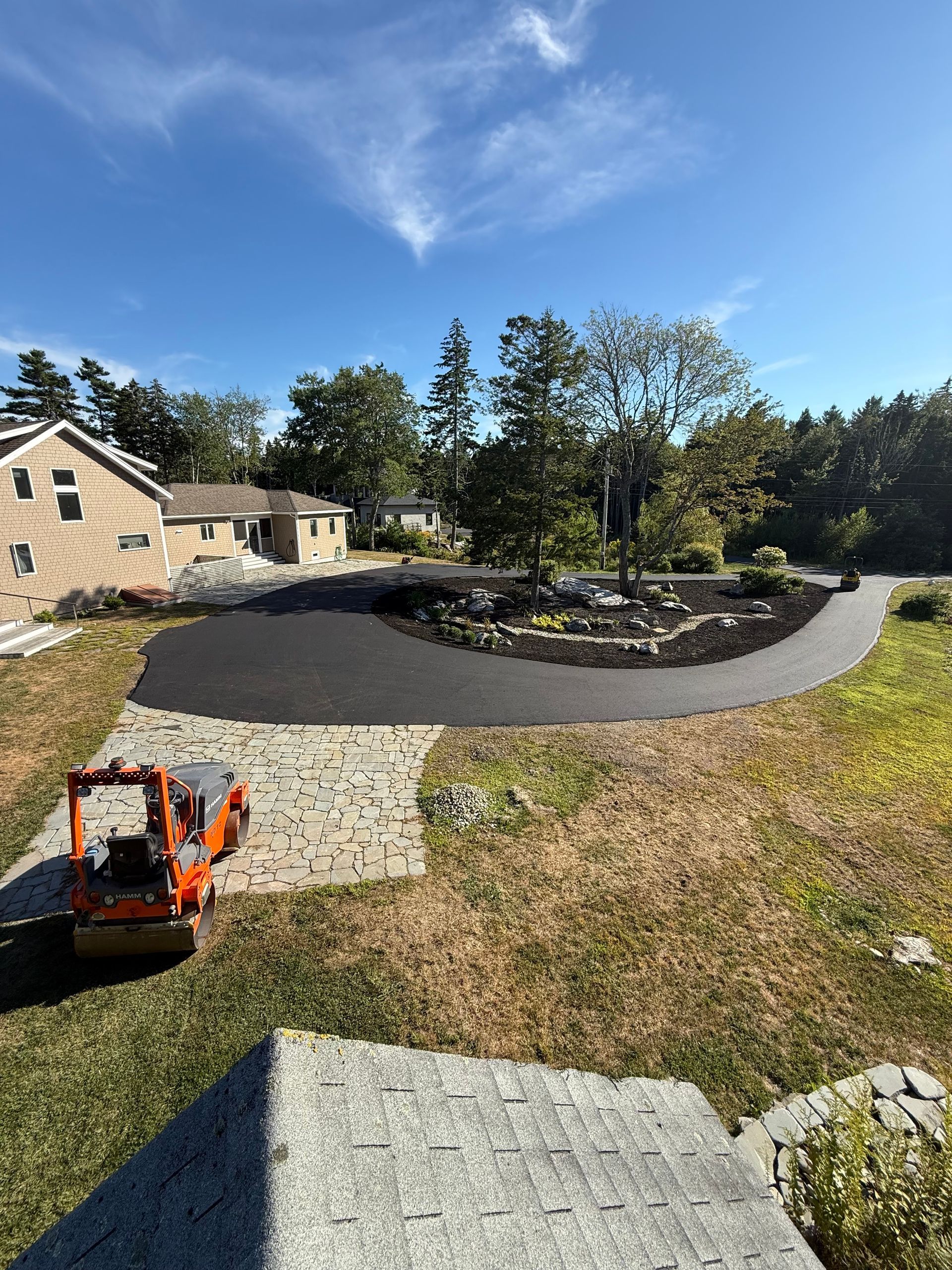 Asphalt driveway curves around a landscaped area with trees, under a blue sky.