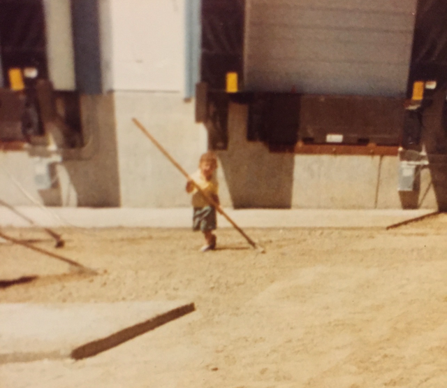A small child in a yellow shirt stands on dirt near industrial loading docks, holding a long wooden pole.